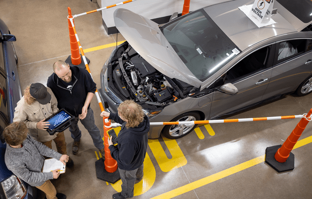 Aerial view of a teacher and 3 students standing in front of an electric vehicle with a barricade around it